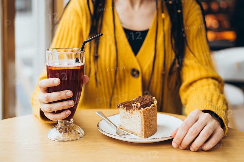 A close-up of a person in a yellow sweater holding a glass with a drink, and a plate with a slice of cake, in a cozy cafe setting Perfect for showcasing relaxation