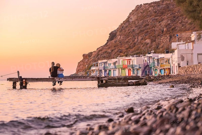 A couple seated on a wooden dock overlooks the vibrant, colorful houses of Klima, Milos Island, during a stunning sunset, creating a serene and romantic atmosphere