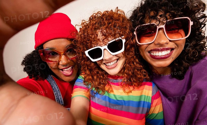 Three multiethnic women in vibrant outfits and sunglasses joyfully pose for a selfie. Their cheerful expressions and bold fashion choices highlight a fun and lively atmosphere