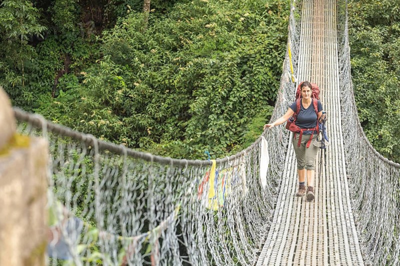 A woman trekker with a backpack crosses a suspension bridge surrounded by lush greenery in the Everest region. She confidently navigates the path, embodying adventure and exploration