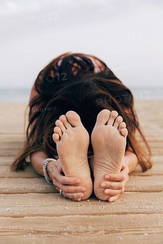 Unrecognizable woman practicing yoga on a beach. The yoga pose shown is a close-up of the Seated Forward Fold (Paschimottanasana) focusing on her feet