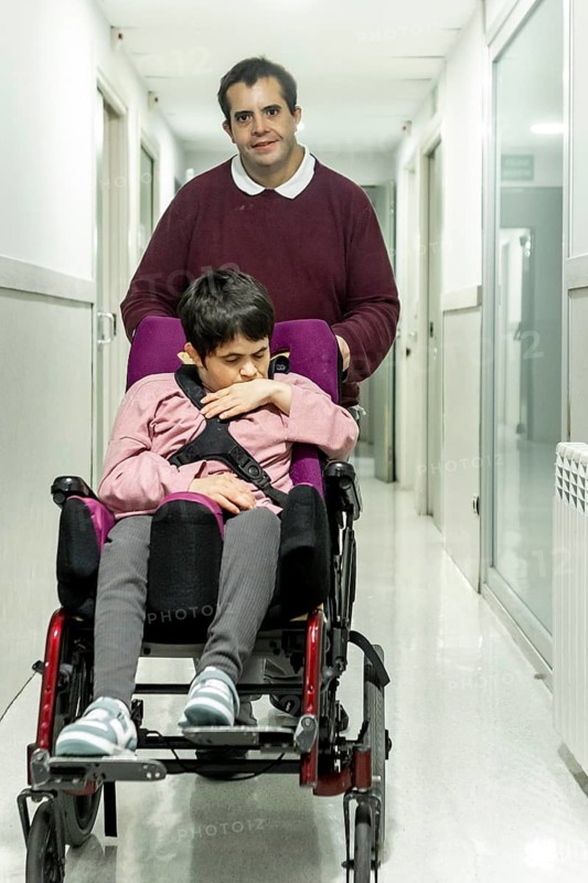 A man assists a woman in a wheelchair down a hallway in a mental health residence. The setting is calm and supportive, emphasizing care and compassion