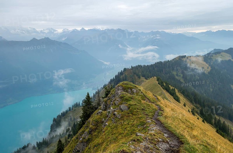 A breathtaking early morning view from a high mountain trail overlooking a turquoise lake surrounded by the rugged peaks of the Bernese Alps, with layers of mist in the valleys