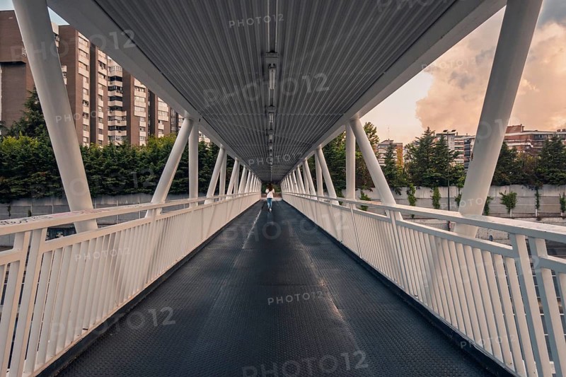 Back view of unrecognizable woman walking on a modern pedestrian bridge in Madrid. The sky is dramatic with storm clouds and orange sunset hues, contrasting the city's urban scenery