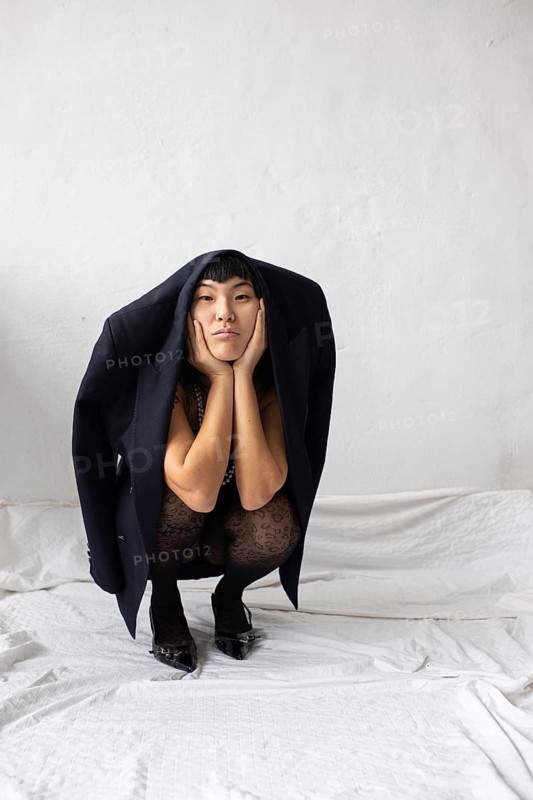 A thoughtful Asian woman poses creatively, with a jacket draped over her shoulders, against a minimalist background. The scene exudes a sense of introspection and originality