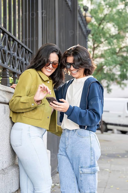 A cheerful couple leans against a fence, laughing and bonding over content on a smartphone. They wear sunglasses, embodying a relaxed and joyful atmosphere in a city setting