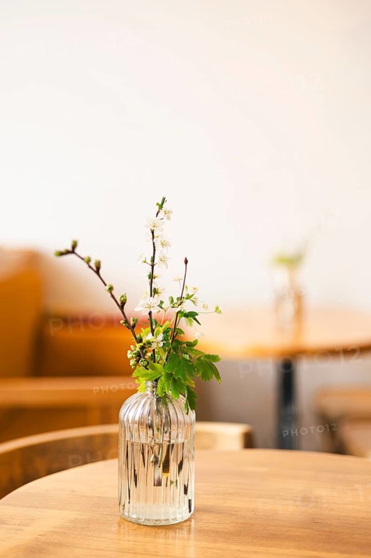 A simple and elegant floral arrangement with delicate white flowers and green leaves sits in a ribbed glass vase on a wooden table, creating a serene and minimalist ambiance in a cafe