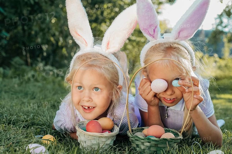Two children with bunny ears delight in an Easter egg hunt outdoors. They smile and playfully hold colorful eggs in wicker baskets on a sunny day, embodying festive joy