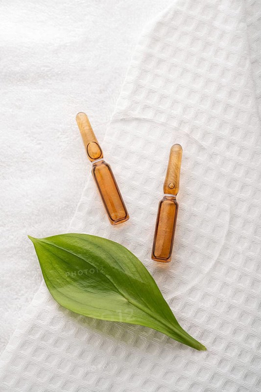 Two brown cosmetic ampoules with anti-aging serum arranged on a textured white towel, accented by a fresh green leaf