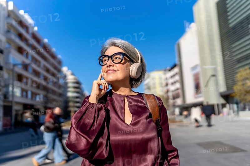 Mature woman with gray hair and glasses smiling contentedly while listening to music with headphones, walking through a vibrant city street on a sunny day