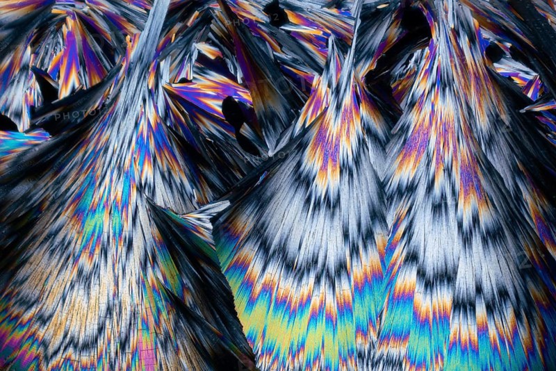 This micrograph showcases stunning hippuric acid crystals, featuring sharp peaks and vibrant colors that include blue, purple, orange, and yellow