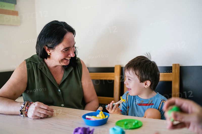 Joyful moment of a mother and her son with Down syndrome playing with pretend food at a table. They smile and engage in a playful activity that fosters connection and learning