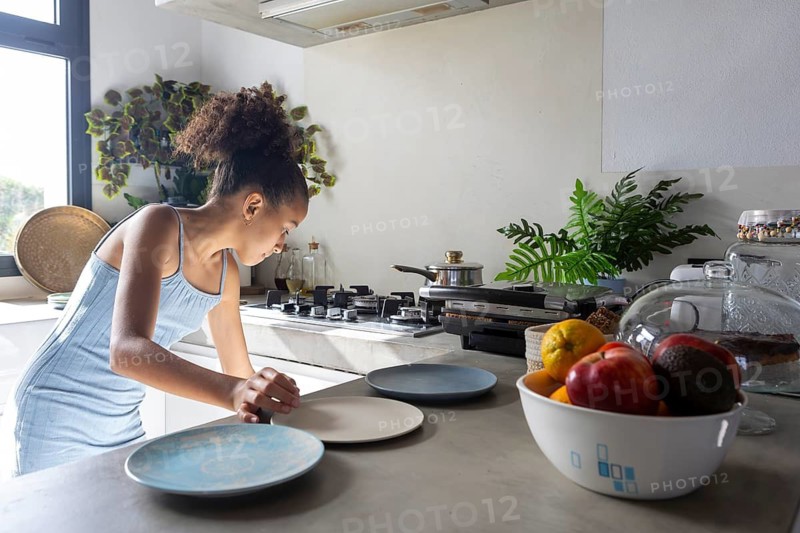 A young girl carefully arranges plates on the kitchen counter, preparing for a meal. The bright, modern kitchen is filled with natural light, fresh fruit, and greenery, creating a cozy atmosphere