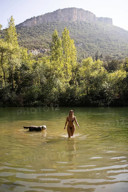 A woman and her dog enjoy a serene day at a lush green lake, surrounded by trees and a rocky hill in the background. The cheerful scene captures the essence of summer days spent in nature