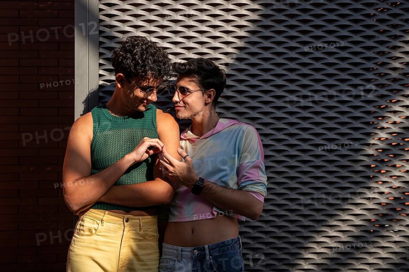 A joyful gay couple shares a warm, sunny moment against a textured urban wall. They are stylishly dressed and radiate affection, symbolizing love and happiness in diversity