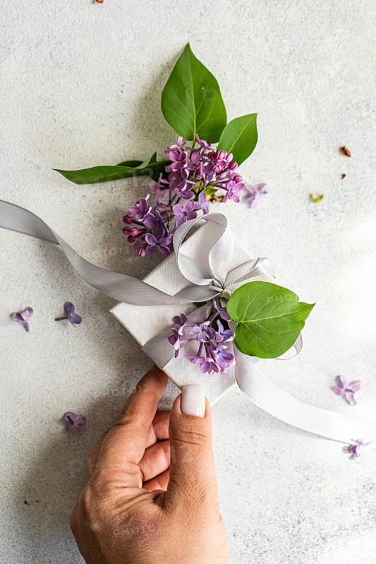 A delicate gift box adorned with lilac flowers and green leaves, tied with a soft white ribbon. A hand gracefully presents the elegant package on a light surface