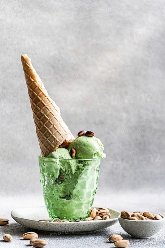 A refreshing scoop of pistachio ice cream with nuts in a textured glass, topped with a crisp waffle cone and garnished with whole pistachios, set on a simple ceramic plate