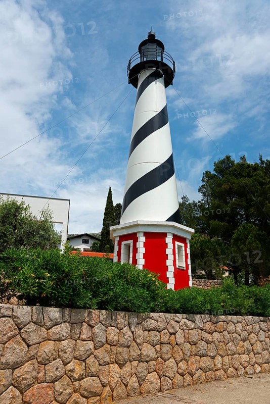 A black and white lighthouse in front of a stone wall, framed by green plants under a clear sky, Starigrad, Starigrad-Paklenica, Zadar, Croatia