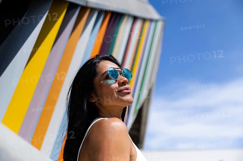 A stylish woman wearing a colorful outfit poses confidently against a backdrop of modern architecture. Her look contrasts with the vibrant urban lines and sky