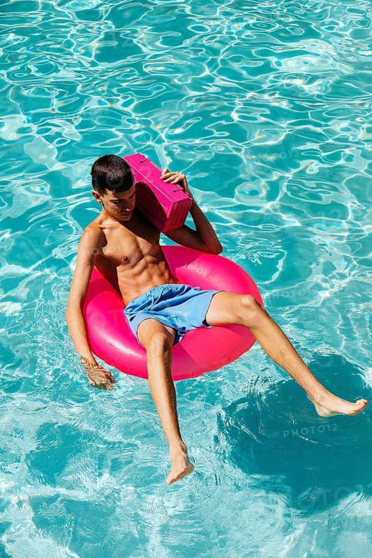 A teenage boy lounges on a bright pink pool float in clear blue water, holding a vibrant pink boombox. They wear light blue swim trunks, soaking up the summer sunshine