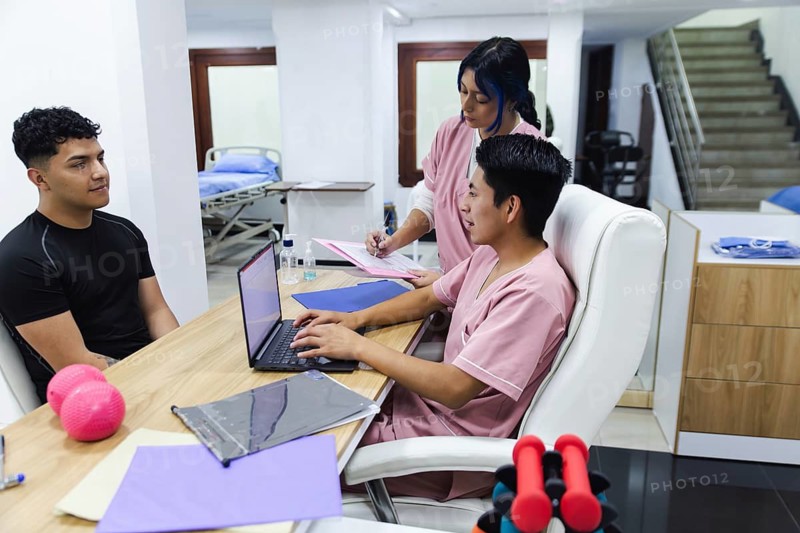 A physiotherapy session in an Ecuadorian clinic. A patient consults with two professionals in scrubs at a desk, highlighting the rehabilitative environment