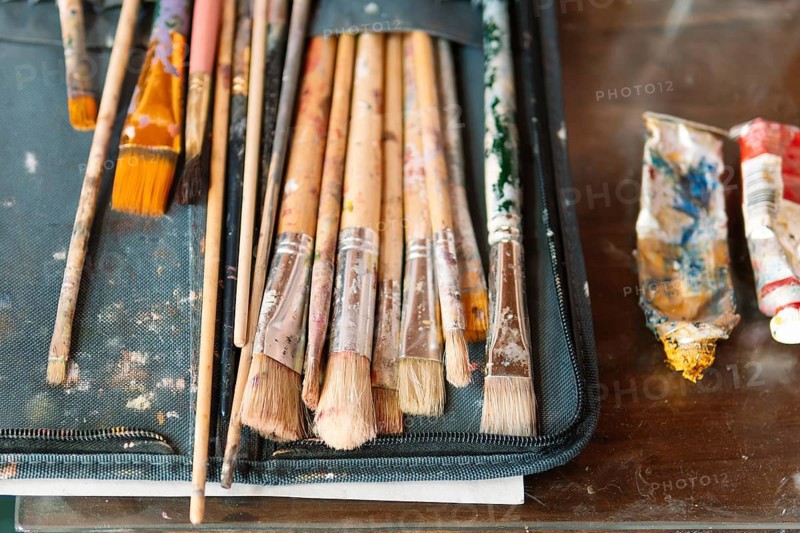 A collection of well-used paintbrushes with dried paint rests in a fabric case, alongside squeezed paint tubes. The worn textures and colors reflect the creativity of an artist's workspace