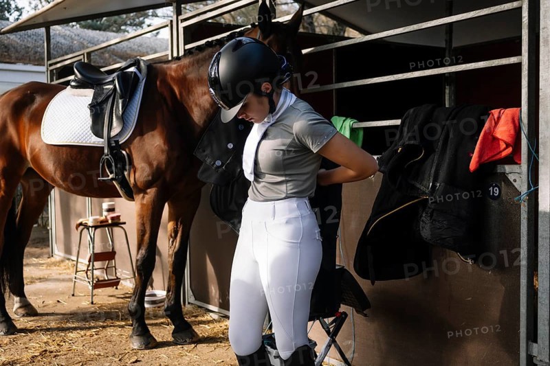 An equestrian, dressed in a helmet and traditional attire, gets ready for a classical dressage event next to a horse in the stable The scene captures the elegance and focus of the sport