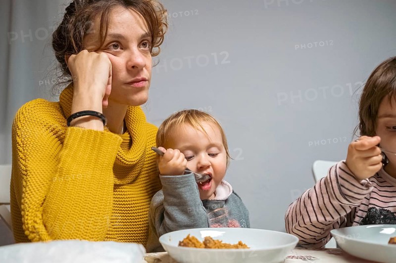 A tired mother sits at the table during a family mealtime, reflecting the challenges of parenting. Her young children eat eagerly, showcasing the demanding yet rewarding aspects of family life
