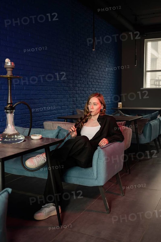 A woman relaxes in a cozy hookah lounge, enjoying the atmosphere. The modern decor features a blue brick wall and soft lighting, enhancing the tranquil ambiance