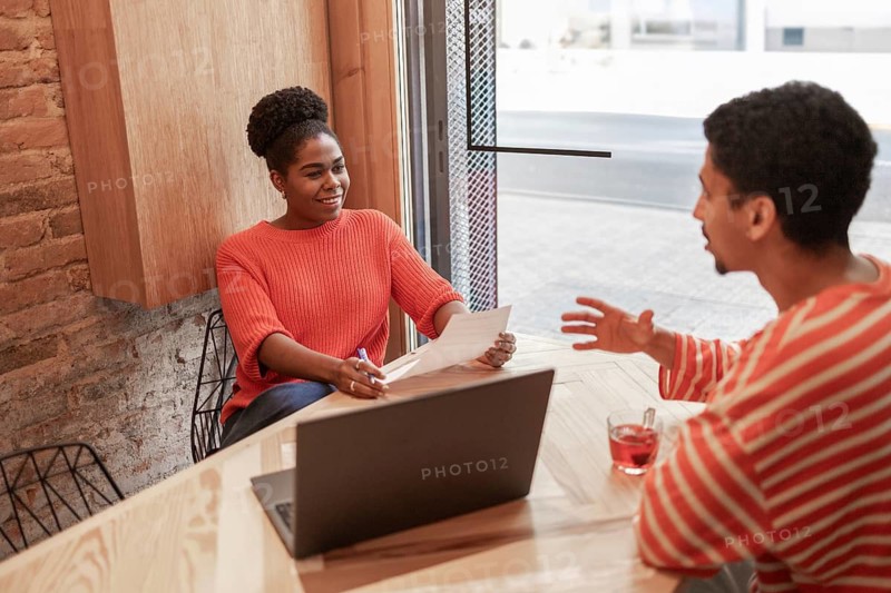 Two people have a discussion in a modern coworking space, sharing ideas with documents and a laptop on the table, promoting collaboration and creative teamwork