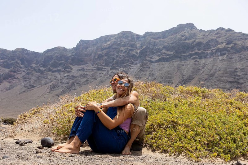 A lesbian couple sits barefoot on rocky ground, surrounded by greenery, embracing and smiling, with a majestic mountainous backdrop under a clear sky, conveying a sense of adventure