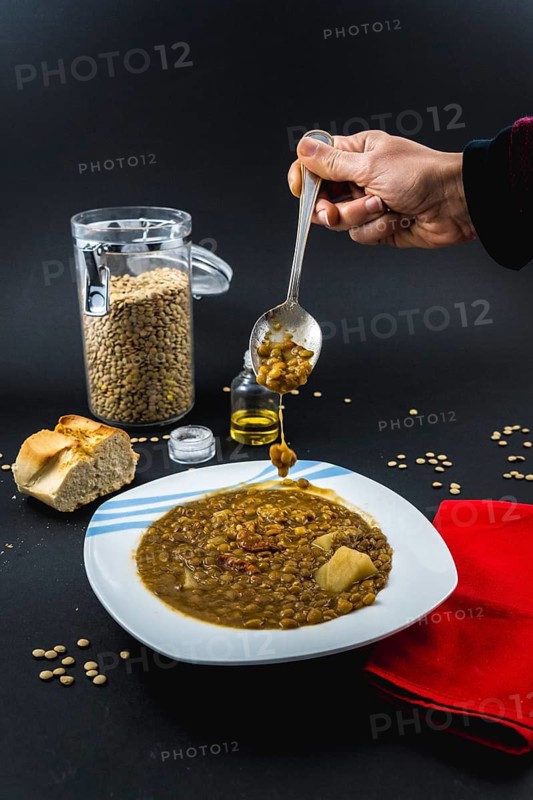A hand of a young woman eating with a spoon inside a plate of Spanish cooked lentils, along with ingredients