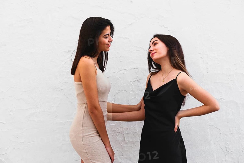 Young sisters, dressed elegantly in white and black dresses, share a joyful moment against a white textured wall, highlighting style