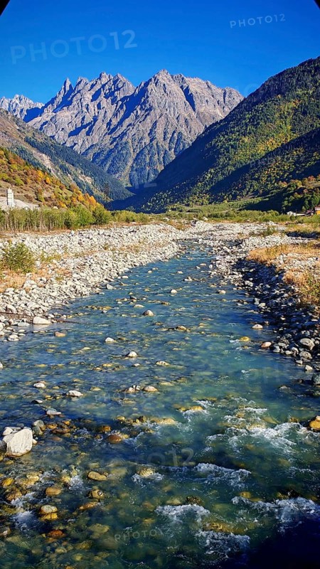 A clear river snakes through a rocky valley surrounded by high, forested mountains under clear skies, Mestia, Chalaadi Glacier, Svaneti, Georgia