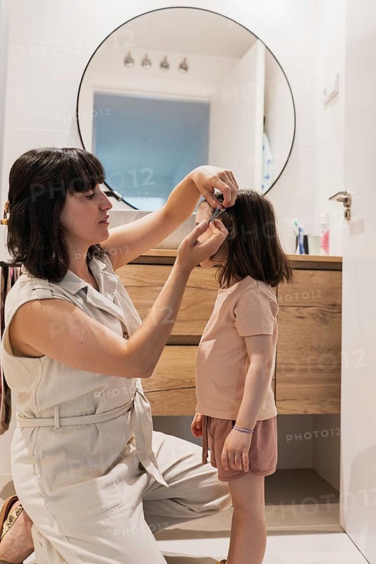 A loving mother gently fixes her young daughter's hair in a cozy, modern bathroom. The warm interaction captures a nurturing family moment, reflecting care and affection
