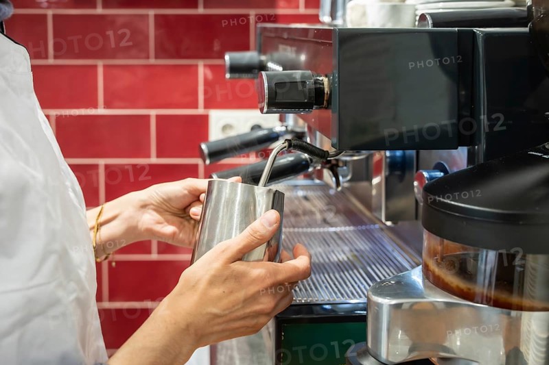 Barista hands holding a stainless steel pitcher, steaming milk using an espresso machine's wand, preparing coffee beverages in a red tiled cafe setting