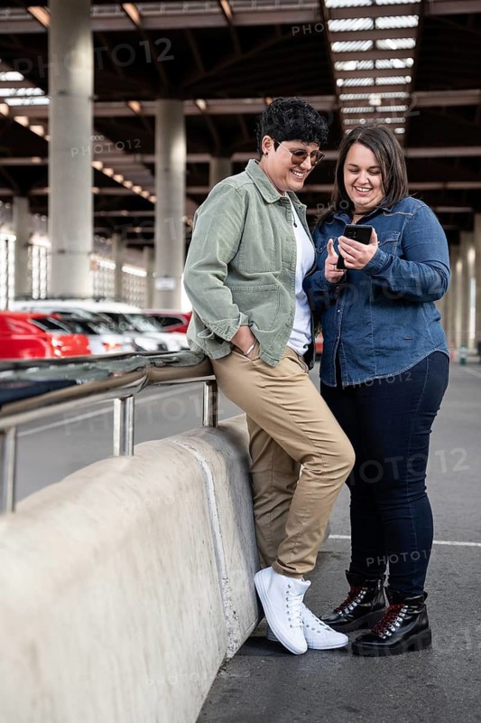 A joyful lesbian curvy couple shares a fun moment while looking at a smartphone. They are standing together in an outdoor setting, embodying love and happiness