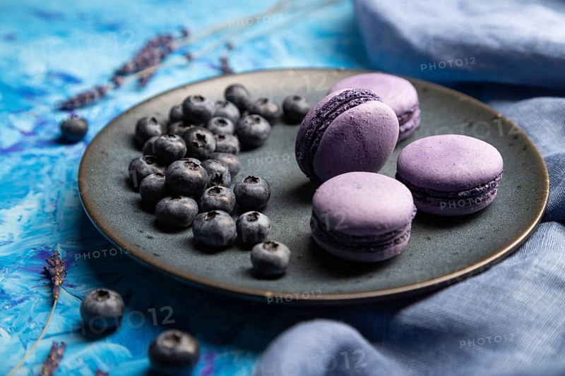 Purple macarons or macaroons cakes with blueberries on ceramic plate on a blue concrete background and blue textile. Side view, close up, selective focus
