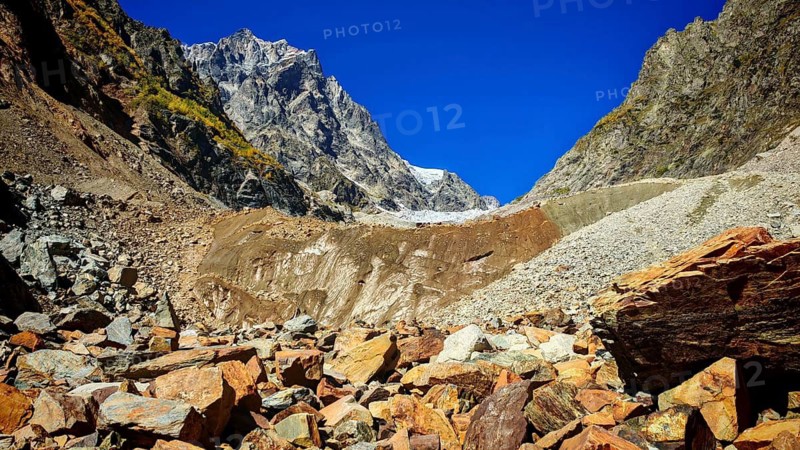 Red debris leads to steep mountain slopes under clear blue skies, hiking to Chalaadi Glacier, Mestia, Svaneti, Georgia