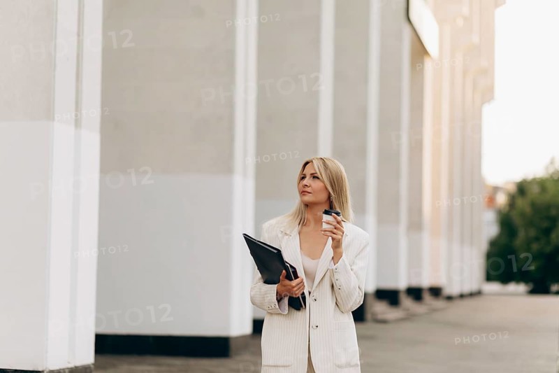 A businesswoman in a stylish white suit holds a coffee cup and folder, standing outdoors. The modern architecture and natural light enhance the urban professional scene
