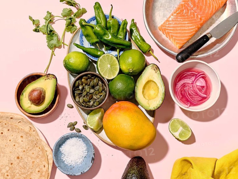Top view of taco ingredients arranged neatly on a pink surface for a culinary concept. Items include fresh salmon, avocados, limes, mango, pepper, salt, coriander, green hot peppers, and tortillas