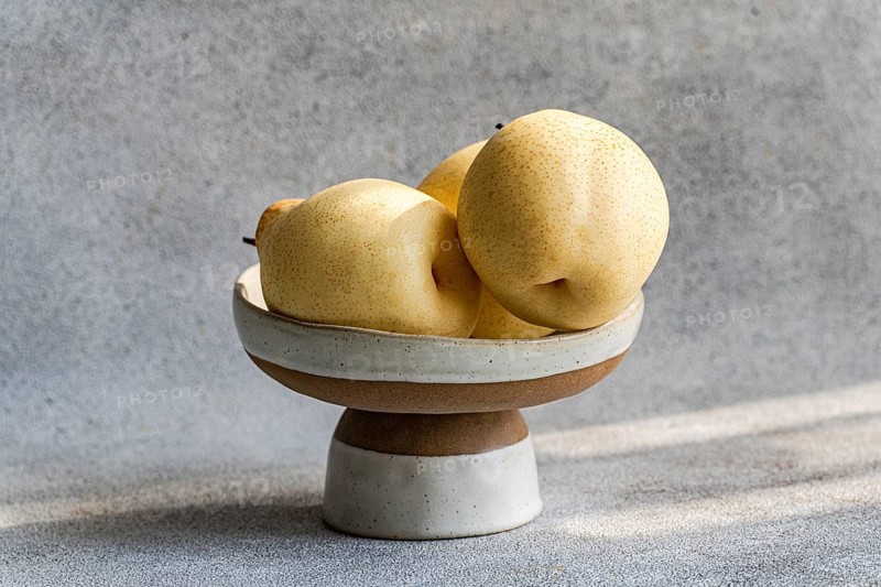 Three Chinese white pears are elegantly displayed in a ceramic bowl against a textured background. The soft light highlights their delicate texture, evoking freshness and simplicity