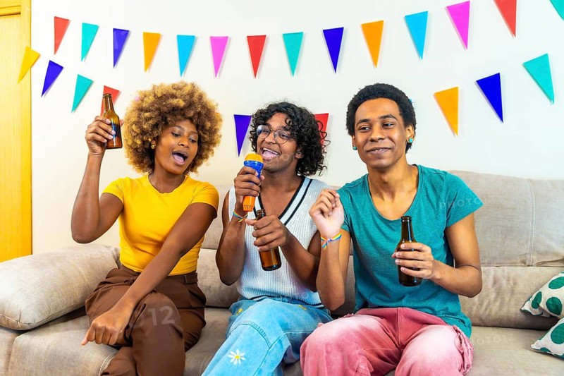 Diverse friends singing and drinking beer during home karaoke sitting on the sofa with party multicolored garlands on the wall