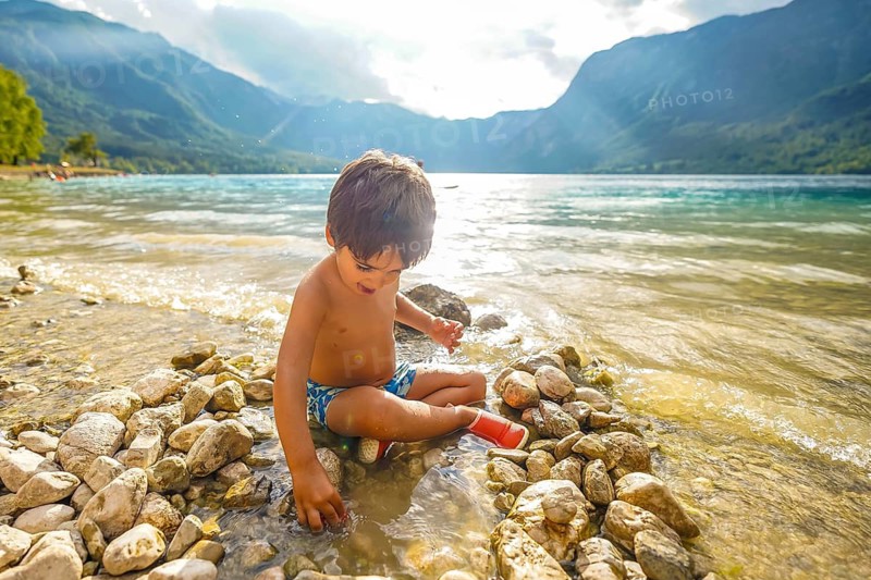 Young boy enjoying summer holidays, playing on the rocky shore of lake bohinj in slovenia, surrounded by beautiful mountain scenery