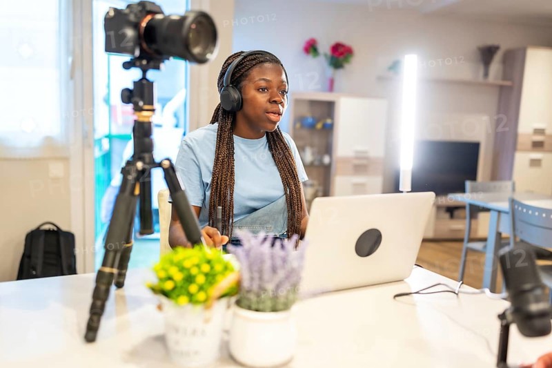 Young woman with headphones using laptop and microphone recording a vlog at home using a camera on a tripod