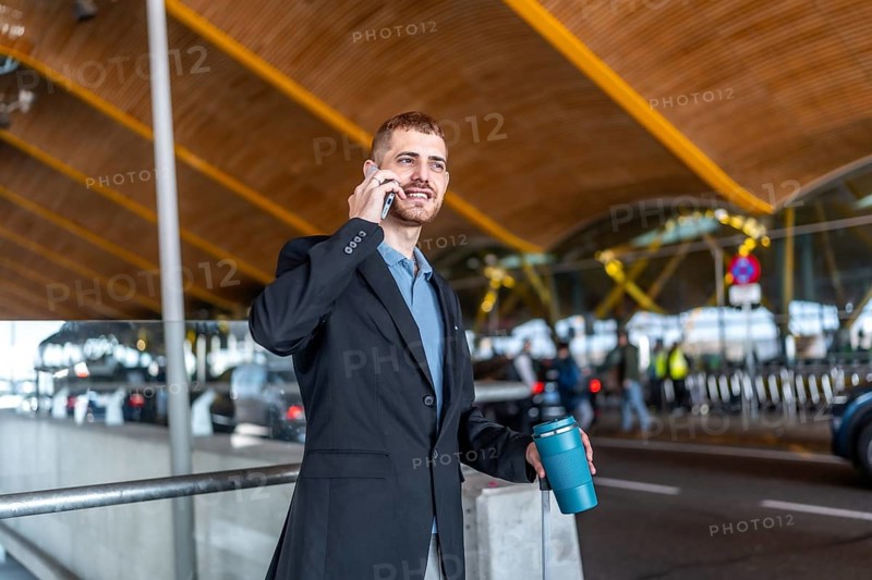 Businessman holding reusable coffee cup and trolley suitcase handle while talking on smartphone at airport