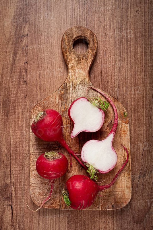 A wooden cutting board features freshly picked radishes, showcasing their vibrant red skins alongside sliced pieces revealing white flesh. The rustic background adds warmth to the composition