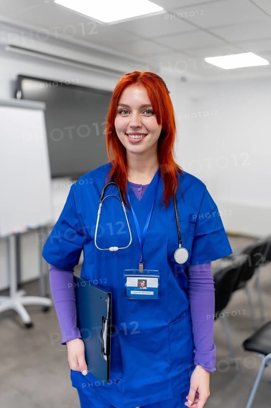 Young woman with red hair wearing blue scrubs and stethoscope, holding a clipboard and id badge, standing in a clinic setting, symbolizing medical care and professionalism