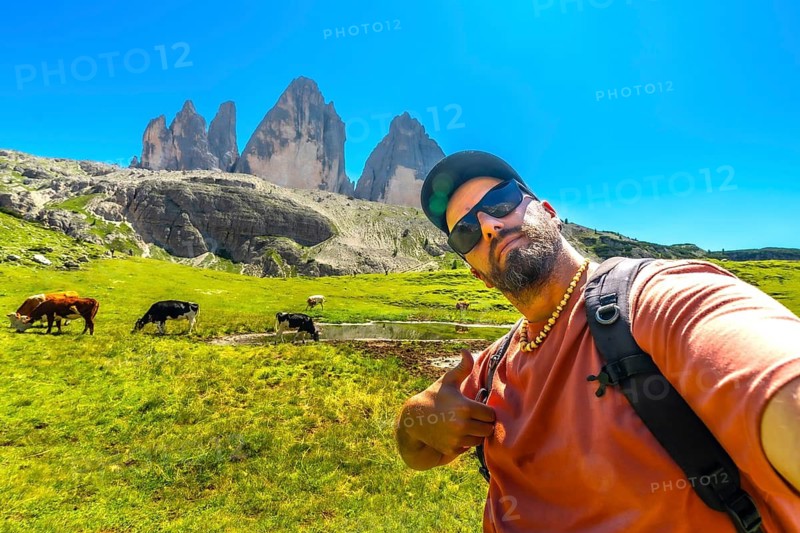Bearded tourist taking a selfie while hiking in the tre cime di lavaredo natural park with cows grazing in the background