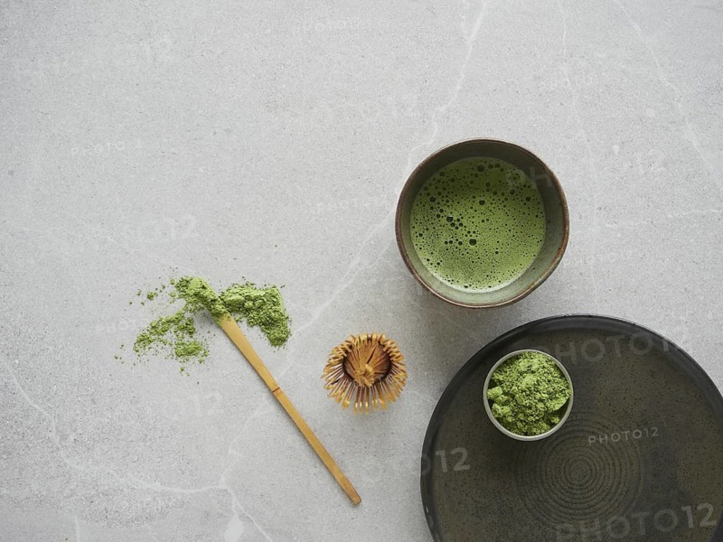 Matcha green tea drink in a bowl, tea powder in bowl and on traditional bamboo spoon next to whisk on gray stone table